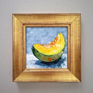 May include: A framed oil painting of a cantaloupe slice. The melon is depicted with a vibrant yellow-orange flesh and a green rind. The artwork is set against a blue background and framed in a gold-colored wooden frame.