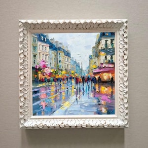May include: Square, white framed oil painting of a city street scene. The artwork depicts a rainy day with reflections on the wet pavement. Buildings line the street, and people are walking.