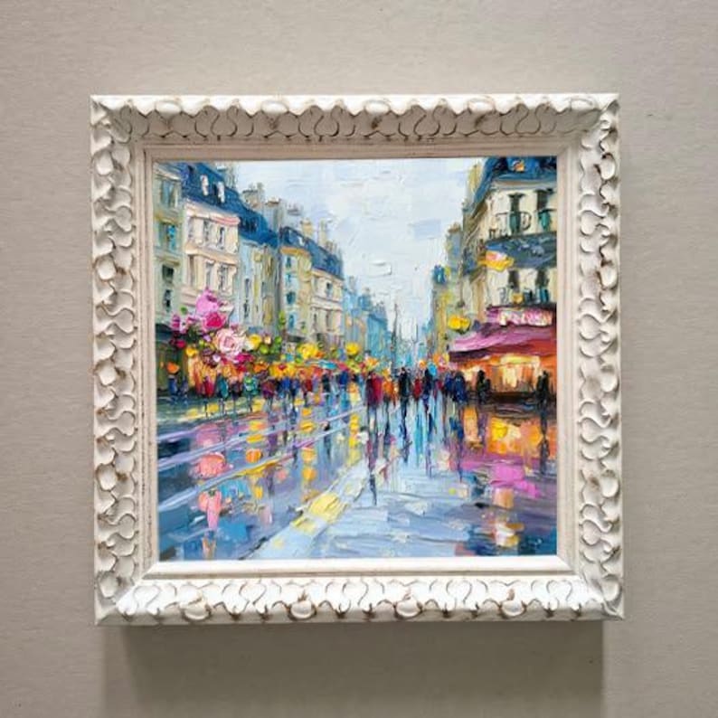 May include: Square, white framed oil painting of a city street scene. The artwork depicts a rainy day with reflections on the wet pavement. Buildings line the street, and people are walking.