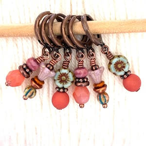 May include: A collection of handcrafted stitch markers. Each marker features a copper-colored ring with decorative beads in shades of coral, pink, and teal. The beads include floral and round shapes, strung on copper wire. The markers are displayed on a wooden knitting needle.