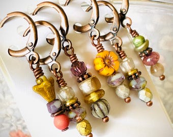 Puede incluir: Primer plano de cinco pendientes hechos a mano con cierres de palanca de color cobre. Cada pendiente presenta una combinación única de cuentas coloridas, incluyendo amarillo, morado y verde, con una variedad de formas y texturas. Los pendientes se exhiben en una tarjeta blanca.
