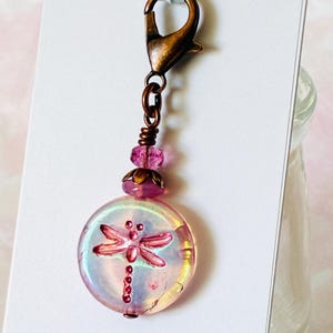May include: A charm featuring a round, iridescent glass bead with a pink dragonfly graphic. The bead is accented with a smaller pink bead and a bronze-toned metal cap, all suspended from a bronze-toned lobster clasp attached to a white card.