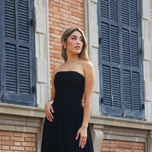 May include: A woman in a black strapless dress stands on a set of stone steps in front of a brick building with closed shutters. She is wearing black high heels and has her hair pulled back.