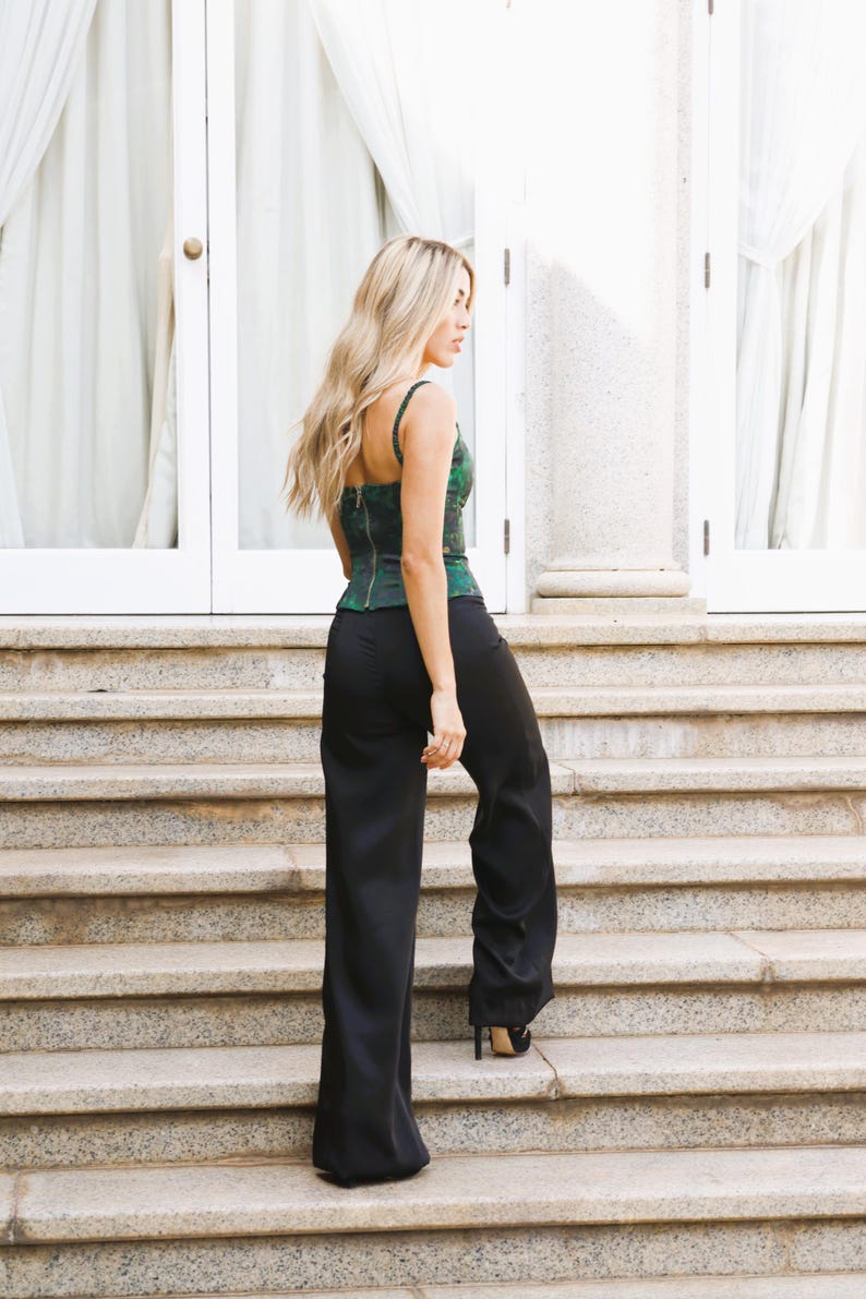May include: A woman wearing a green and black patterned corset top and black wide-leg pants ascends a stone staircase. The top has thin straps and a zipper in the back. The pants are long and flowing. The background includes white doors and curtains.