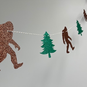 May include: A brown glitter Bigfoot and green glitter pine tree garland. The garland is strung with white twine.