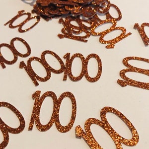 Puede incluir: Confetti de purpurina de cobre con el número 100 impreso en cada pieza. El confeti está esparcido sobre una superficie blanca.