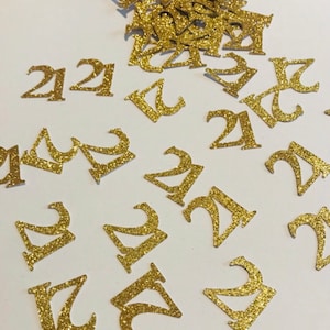 May include: Gold glitter confetti with the number 21 printed on each piece. The confetti is scattered on a white surface.