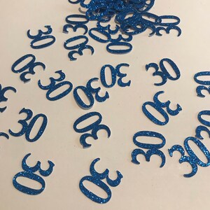 May include: Blue glitter confetti with the number 30 printed on each piece. The confetti is scattered on a white surface.