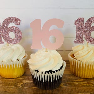 Puede incluir: Tres cupcakes decorados con toppers de cupcake "16" de purpurina rosa. Dos cupcakes tienen glaseado blanco y uno tiene glaseado de chocolate.