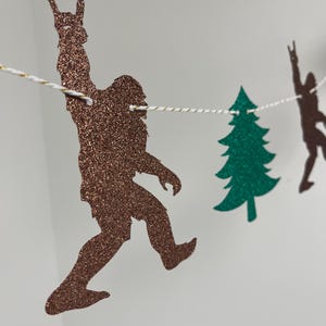 May include: A brown glitter Bigfoot, a green glitter pine tree, and a brown glitter Bigfoot with arms raised in the air are strung together on a white string. The figures are cutouts and are part of a banner.