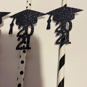 Puede incluir: Pajitas rayadas en blanco y negro y con lunares con gorros de graduaci&oacute;n de purpurina negra y el a&ntilde;o 2021 adjunto.