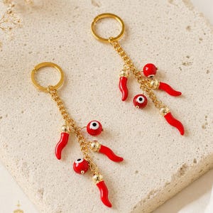 May include: Two gold keyrings with dangling charms. Each keyring features a gold chain with red chilli pepper charms, red bead charms with an evil eye design, and gold accents. The keyrings are displayed on a light-coloured surface.