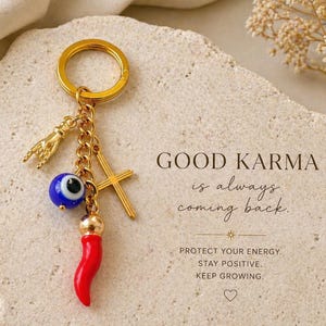 May include: Gold-coloured keychain with charms including a hand gesture, a blue evil eye, a red chilli pepper, and a cross. The text "GOOD KARMA is always coming back" is displayed on a textured background. The brand name "GOOD KARMA AVENUE" is also visible.