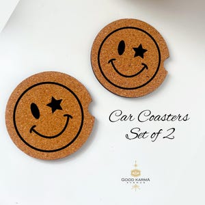 May include: Two round car coasters with a smiley face design. The smiley faces have a star for one eye and a curved line for the mouth. The coasters are made of a light brown material and have a cutout for a car cup holder.