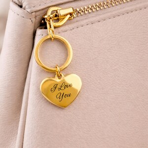 May include: A gold heart-shaped keyring inscribed with "I Love You" hangs from a gold ring attached to a light-coloured purse. The purse has a gold zip and hardware. The keyring is a romantic accessory.