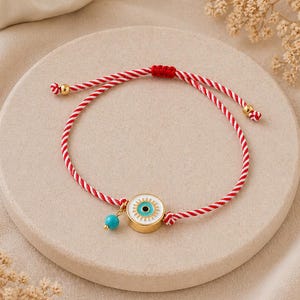 May include: A red and white cord bracelet with a gold-tone eye charm and a turquoise bead. The bracelet has adjustable knots and is displayed on a neutral surface. The words "GOOD KARMA" are at the bottom of the image.