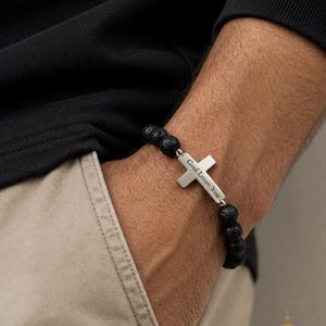 May include: Black lava stone bead bracelet with a silver cross pendant. The cross is engraved with the words "God Loves You". The bracelet is worn on a wrist.