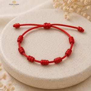 May include: A red string bracelet with multiple knots. The bracelet is displayed on a light-colored, round, stone-like surface. The bracelet has an adjustable sliding knot closure.