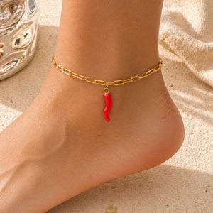 May include: Gold-coloured chain anklet featuring a red chilli pepper charm. The anklet is worn on an ankle. The charm hangs down. The background is a sandy surface. The text "GOOD KARMA AVENUE" is visible.
