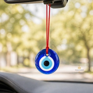 May include: A blue and white Evil Eye car charm suspended from a red string, hanging from a car's rearview mirror. The charm features a circular design with a blue background, white and light blue concentric circles, and a black centre. The background is blurred.