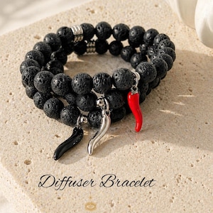 May include: Black lava stone diffuser bracelet with three charm pendants: a black chili pepper, a silver chili pepper, and a red chili pepper. The bracelet is displayed on a textured surface. The text "Diffuser Bracelet" is visible.