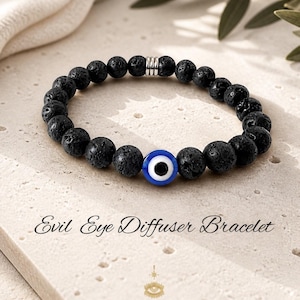 Puede incluir: Pulsera de cuentas de piedra de lava negra con un dije de ojo turco azul y blanco. La pulsera tiene una cuenta espaciadora plateada. El texto "Evil Eye Diffuser Bracelet" se muestra debajo.