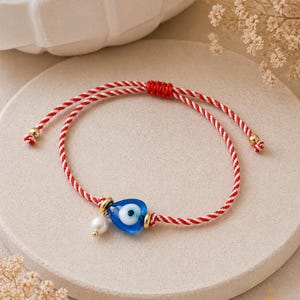 May include: A red and white cord bracelet with a blue evil eye charm and a small white pearl. The charm is surrounded by gold-colored metal. The bracelet is on a neutral-colored surface.
