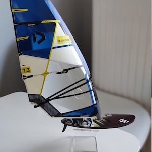 Puede incluir: Una vela de windsurf azul y blanca con correas negras y detalles amarillos. La vela está etiquetada con "Duotone" y "E-Type". La vela está montada en un soporte de acrílico transparente.