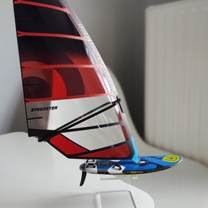 Puede incluir: Una vela de windsurf roja, negra y plateada con la palabra "SPEEDSTER" impresa en ella. La vela está montada en un soporte de acrílico transparente.