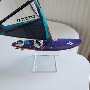 May include: A miniature windsurf board with a purple deck and a sail. The sail is white, teal, and black with the text "DUOTONE". The board has white accents and is displayed on a clear stand.