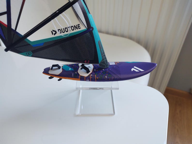 May include: A miniature windsurf board with a purple deck and a sail. The sail is white, teal, and black with the text "DUOTONE". The board has white accents and is displayed on a clear stand.