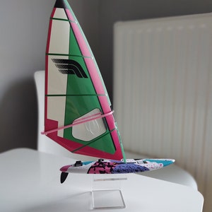 Puede incluir: Un modelo en miniatura de una tabla de windsurf con una vela rosa, verde y blanca. La vela tiene un logotipo negro con un diseño de pájaro estilizado. La tabla está decorada con un patrón abstracto colorido. El modelo se muestra en un soporte de acrílico transparente.