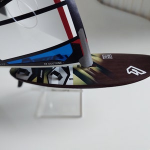 Puede incluir: Primer plano de una tabla de windsurf marr&oacute;n y blanca con un logotipo blanco y la palabra "Duotone" impresa en la vela. La tabla est&aacute; sobre una superficie blanca.