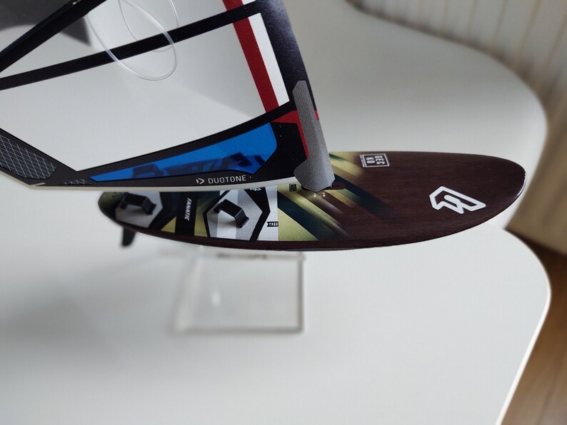 Puede incluir: Primer plano de una tabla de windsurf marr&oacute;n y blanca con un logotipo blanco y la palabra "Duotone" impresa en la vela. La tabla est&aacute; sobre una superficie blanca.