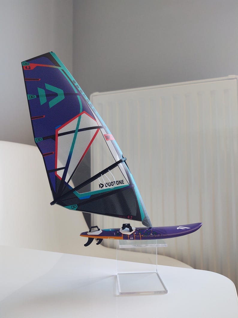 May include: A miniature windsurfing board and sail set. The sail is primarily purple with teal, red, and white accents. The board is purple with orange and white details. The set is displayed on a clear stand.