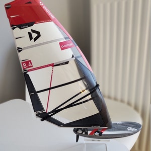 Puede incluir: Una vela de windsurf con un diseño negro, blanco y rojo, con el logotipo "DUOTONE". La vela está etiquetada como "8.4" y "WARP". La vela está montada en una pequeña tabla de windsurf.