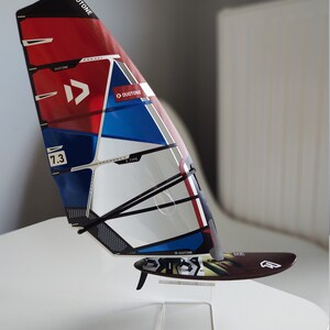 Puede incluir: Una vela de windsurf roja, blanca y azul con el logotipo de Duotone y el texto "E-Type" y "7.3". La vela est&aacute; montada en un soporte de acr&iacute;lico transparente.