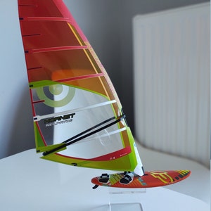 Puede incluir: Una vela de windsurf roja, amarilla y verde con el texto "Hornet Neil Pryde" y "7.2" impreso en ella. La vela está montada en un soporte de acrílico transparente.