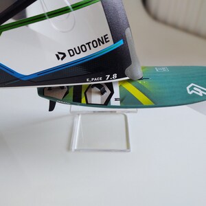 K&ouml;nnte beinhalten: Ein gr&uuml;n-gelbes Windsurfboard mit einem wei&szlig;en Logo und dem Text "E_PACE 7.8" auf dem Deck. Das Board steht auf einem durchsichtigen Acryl-St&auml;nder. Das Board ist teilweise von einem wei&szlig;en und schwarzen Windsurfsegel mit dem Duotone-Logo verdeckt.