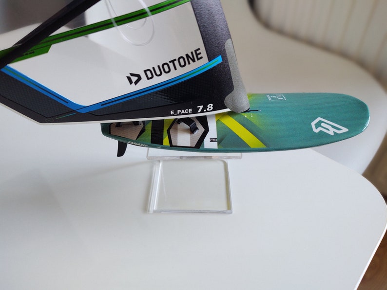 K&ouml;nnte beinhalten: Ein gr&uuml;n-gelbes Windsurfboard mit einem wei&szlig;en Logo und dem Text "E_PACE 7.8" auf dem Deck. Das Board steht auf einem durchsichtigen Acryl-St&auml;nder. Das Board ist teilweise von einem wei&szlig;en und schwarzen Windsurfsegel mit dem Duotone-Logo verdeckt.