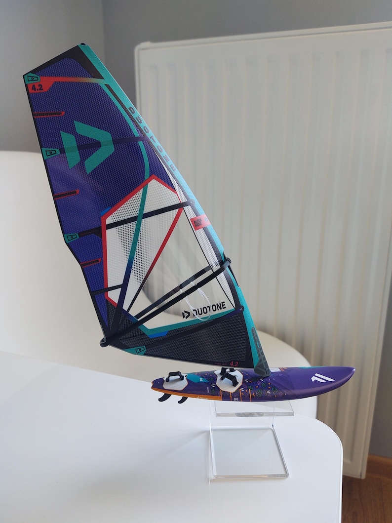 May include: A miniature windsurfing setup featuring a purple and teal sail with the text "DUOTONE" and a matching purple board. The sail has a blue and red design with the number "4.2". The board has black fins.