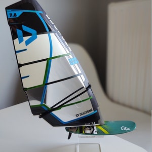K&ouml;nnte beinhalten: Ein schwarzes, wei&szlig;es, blaues und gr&uuml;nes Windsurfsegel mit einem schwarzen Gurt und dem Text "Duotone E-Pace 7.8" auf dem Segel. Das Segel ist auf einem klaren Acrylst&auml;nder montiert.