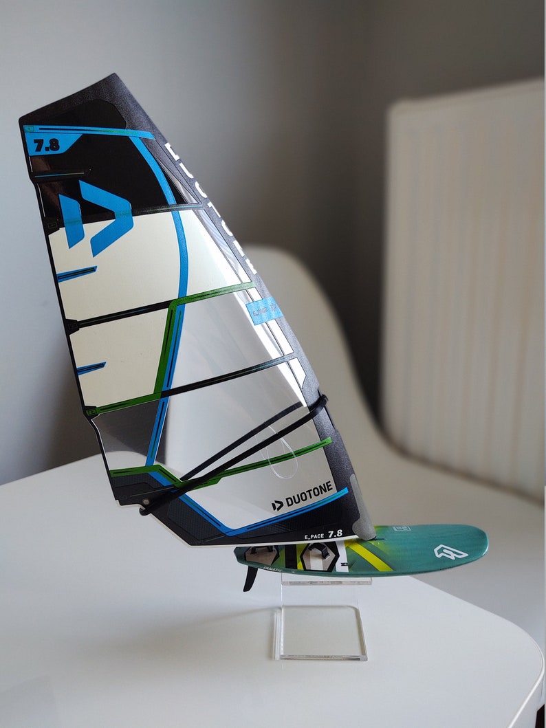 K&ouml;nnte beinhalten: Ein schwarzes, wei&szlig;es, blaues und gr&uuml;nes Windsurfsegel mit einem schwarzen Gurt und dem Text "Duotone E-Pace 7.8" auf dem Segel. Das Segel ist auf einem klaren Acrylst&auml;nder montiert.
