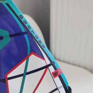 May include: A close-up of a windsurfing sail with the word "DUOTONE" printed in black. The sail features a blue and teal design with red and white accents. A small red rectangle with the words "SUPER HERO" is visible.