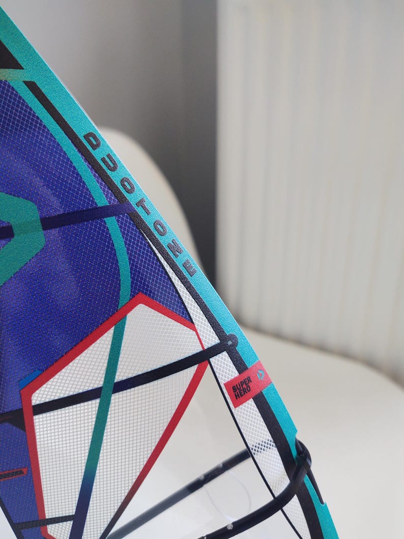 May include: A close-up of a windsurfing sail with the word "DUOTONE" printed in black. The sail features a blue and teal design with red and white accents. A small red rectangle with the words "SUPER HERO" is visible.
