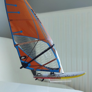Puede incluir: Un modelo de vela y tabla de windsurf. La vela es naranja, azul y blanca con detalles negros. La tabla es blanca con un patrón negro y gris. La vela está montada en un soporte de acrílico transparente.