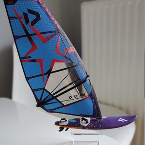 May include: A blue and black windsurf sail with a red star design and the text "Duotone" and "Super Star 4.2". The sail is mounted on a purple surfboard with a colorful design.
