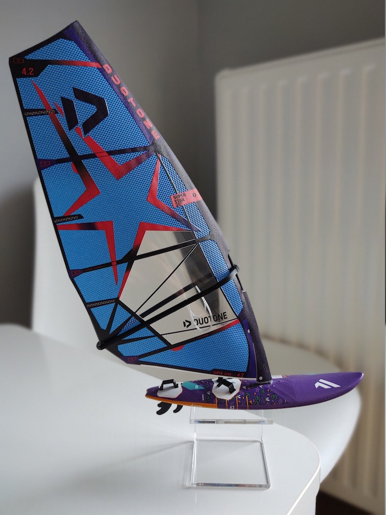 May include: A blue and black windsurf sail with a red star design and the text "Duotone" and "Super Star 4.2". The sail is mounted on a purple surfboard with a colorful design.