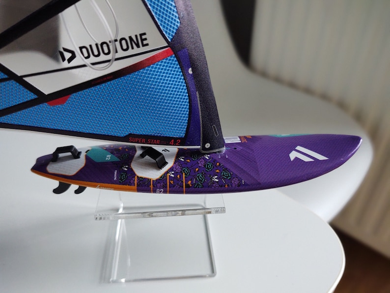 May include: A purple and white windsurf sail with a black and blue DUOTONE logo and the text "SUPER STAR 4.2" on the sail. The sail is on a clear acrylic stand.