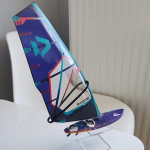 May include: A miniature windsurfing sail and board set. The sail is primarily blue with teal and red accents, and the board is purple. The sail features the text "DUOTONE" and the number "4.2". The set is displayed on a clear stand.